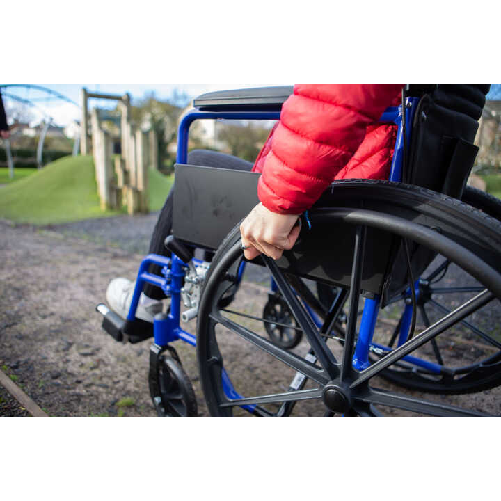 A person in a red jacket is holding the wheel of a blue wheelchair on a playground.