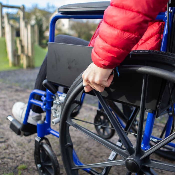 A person in a red jacket is holding the wheel of a blue wheelchair on a playground.
