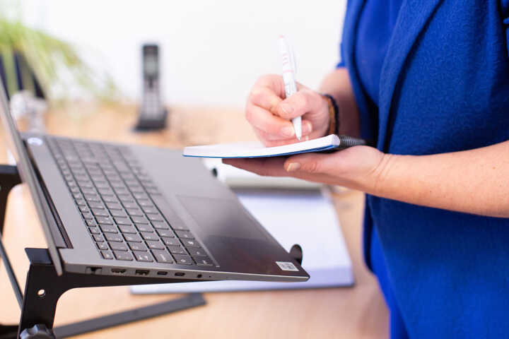 A person in a blue shirt writes notes in a notebook while using a laptop.