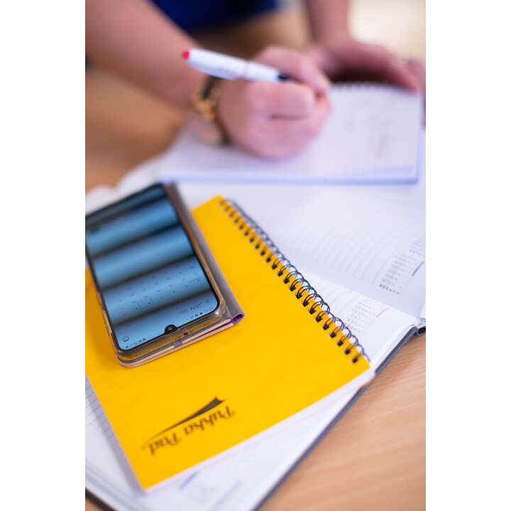 A smartphone rests on a yellow notebook next to a hand writing in a notebook.