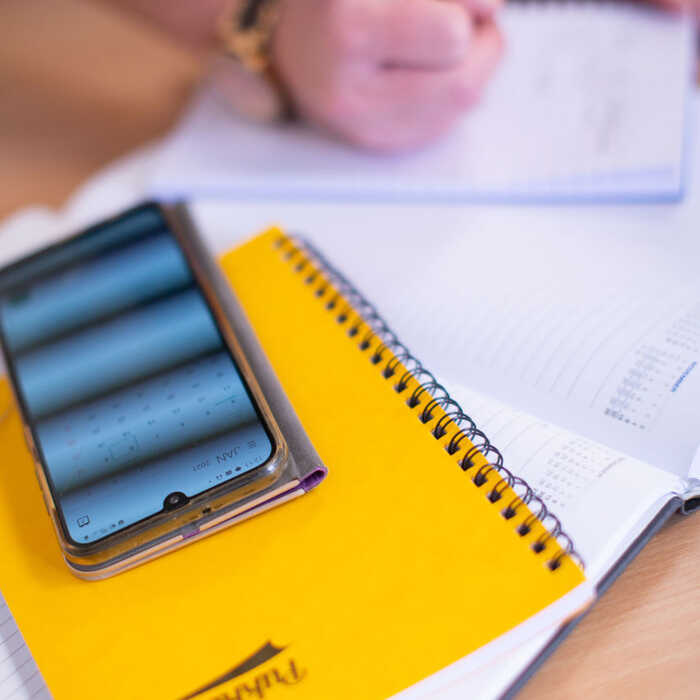 A smartphone rests on a yellow notebook next to a hand writing in a notebook.