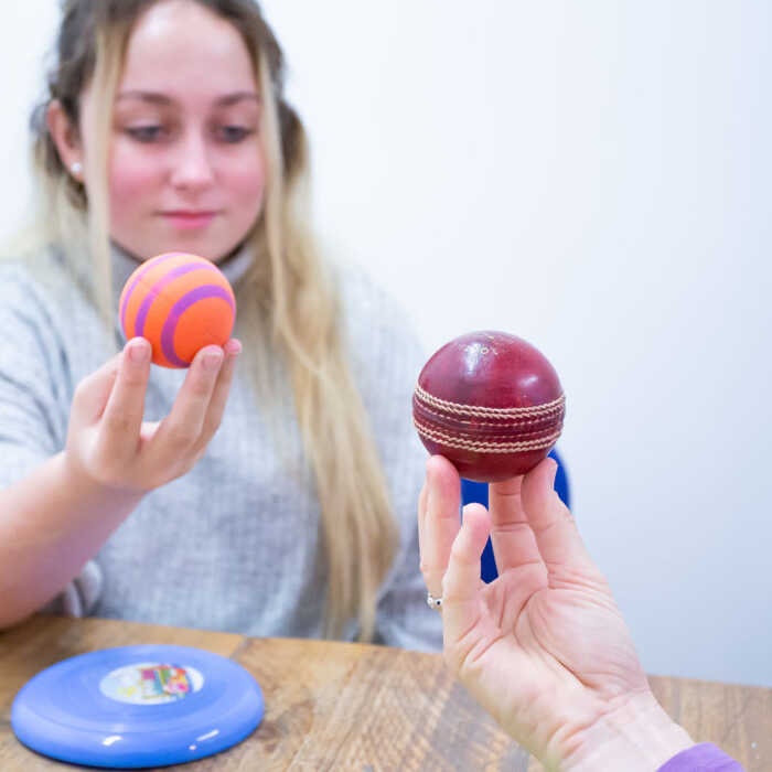 A young woman holds a colorful ball, while another person reaches out with a cricket ball.