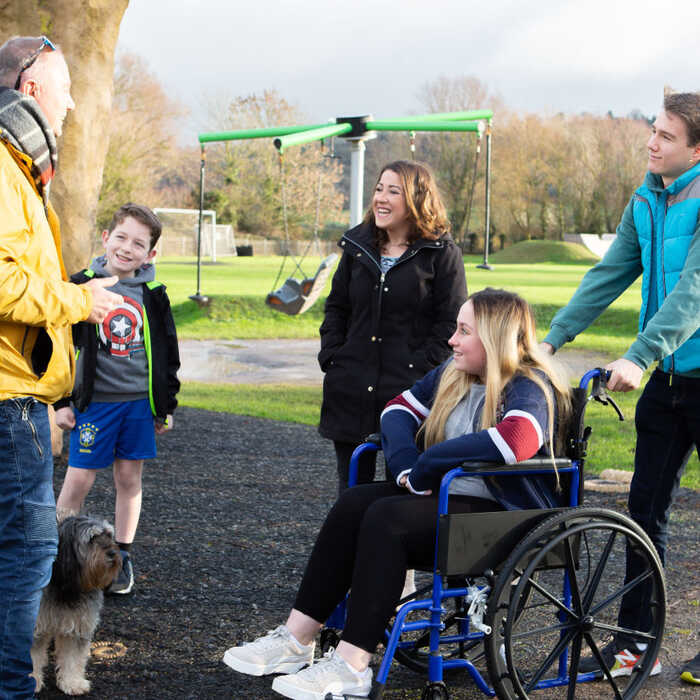 A group of four people interact outdoors, with one person in a wheelchair and a dog nearby.