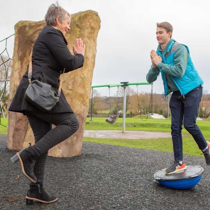 A woman watches as a young man balances on a wobble board in a park.