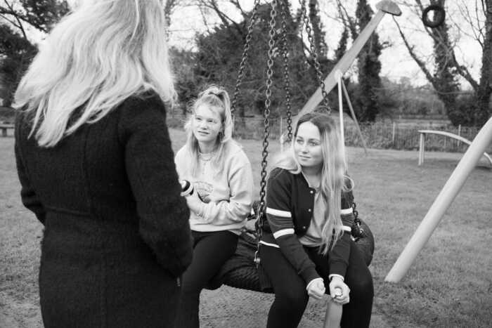 Two girls sit on a swing at a park, talking to a woman standing nearby. Black and white image.